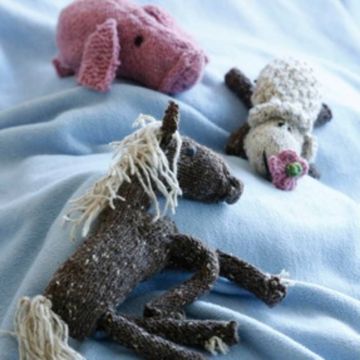 Drei gestrickte Stofftiere liegen auf einer hellblauen Decke: ein rosa Schwein oben links, ein weißes Schaf mit rosa Blume im Maul oben rechts und ein braunes Pferd mit heller Mähne und Schweif unten im Vordergrund. Alle Tiere bestehen aus Wolle und haben