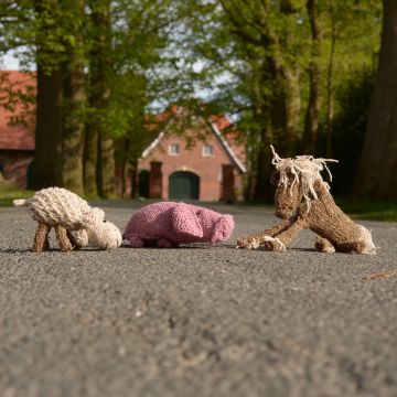 Drei gestrickte Stofftiere liegen auf einer Hofeinfahrt: ein rosa Schwein oben links, ein weißes Schaf mit rosa Blume im Maul oben rechts und ein braunes Pferd mit heller Mähne und Schweif unten im Vordergrund. Alle Tiere bestehen aus Wolle und haben